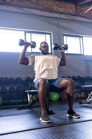 Seated man pressing hex dumbbells in industrial gym wearing headphones and wristwatch