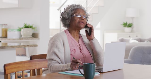 Elderly African American woman working from home. She is sitting at a table with a laptop and notepad while speaking on the phone. There is a coffee cup on the table, emphasizing a busy work environment. Ideal for content about remote work, senior independence, digital literacy among seniors, and balancing professional and personal life while working from home.