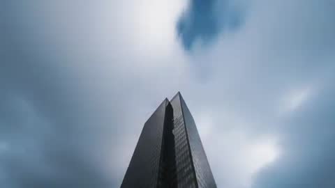 Drone rising past reflective twin-peak skyscraper toward fast-moving moody cloudscape