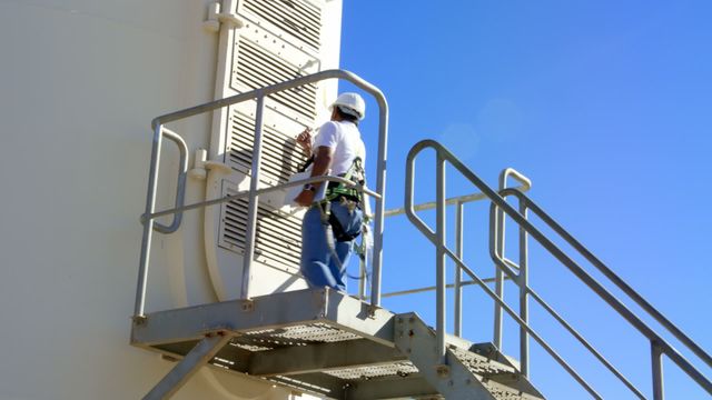 Engineer performs maintenance on wind turbine via metal ladder and platform, despite the sunny, cloudless day. Image suggests use in contexts related to renewable energy, engineering profession, or safety procedures, ideal for illustrating topics on sustainable technology and workforce in energy sectors.