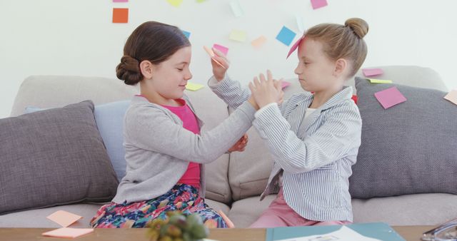 Two young girls on a couch enjoying arts and crafts at home. Colorful sticky notes are visible in the background. Great for concepts related to child development, creative activities for kids, playful learning, and family bonding. Suitable for use in educational content, parenting advice articles, and advertisements for children's art supplies.