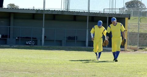 Two baseball teammates walking across sunlit outfield in yellow and blue uniforms carrying mitts