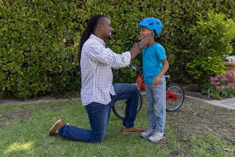 African american father kneeling adjusting blue helmet on young son before bike ride