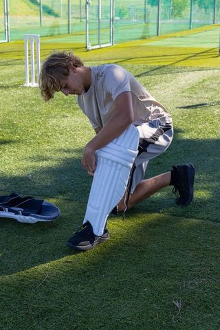 Teen male kneeling fitting white cricket batting pads on turf at practice nets and stumps