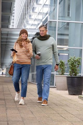 Senior couple walking under glass canopy carrying coffee and phone in modern city plaza