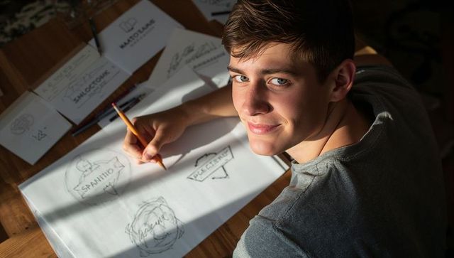 Young designer sketching logo concepts on sunlit wooden desk with pencil and sketchbook