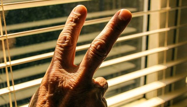 Senior hand making v sign through sunlit venetian blinds casting warm shadow lines