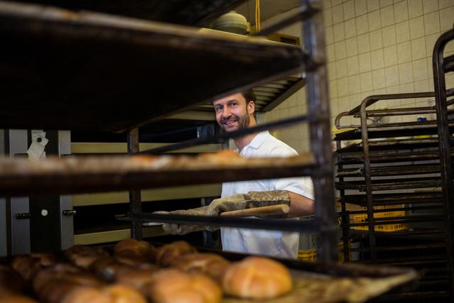 Smiling baker removing fresh buns from the oven in a bakery. Professional chef working in a kitchen, handling freshly baked bread with care. Ideal for illustrating artisanal baking, small business, culinary skills, or the daily routine of a professional baker.