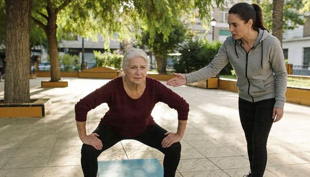 Senior woman performing wide squat in urban park with personal trainer coaching outdoors