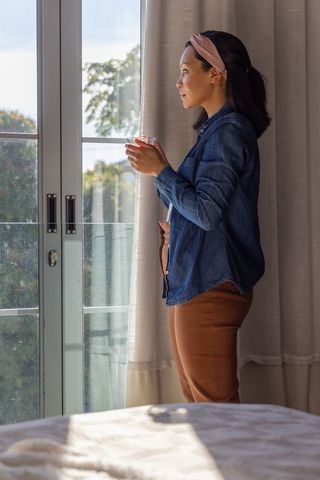 Asian woman standing by glass door holding cup, serene morning sunlight, quiet contemplation