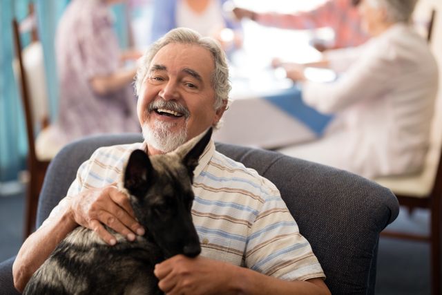 Senior man sitting comfortably with a puppy on his lap, smiling warmly. Background shows other elderly individuals engaging in activities. Ideal for use in advertisements for retirement homes, senior living communities, pet therapy programs, or articles about the benefits of pets for seniors.