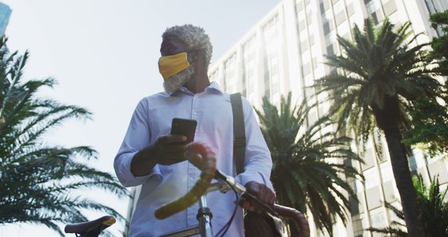 Elderly man wearing yellow face mask, holding smartphone and bicycle. Palm trees and tall buildings in background. Suitable for topics on urban lifestyle, COVID-19 safety measures, fitness, eco-friendly transportation, and senior citizens staying active.