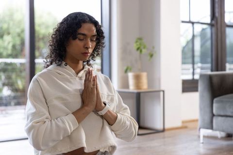 Non-binary adult meditating with hands in prayer in sunlit minimalist living room