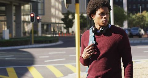 Young man walking urban crosswalk gripping backpack strap with headphones, sunlit commuter