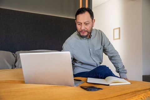Mature asian man working on laptop in modern bedroom home office taking notes on notebook