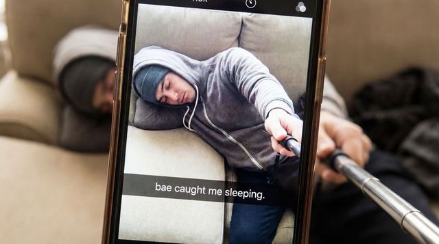 Smartphone selfie showing partner sleeping on sofa in hoodie with selfie stick caption