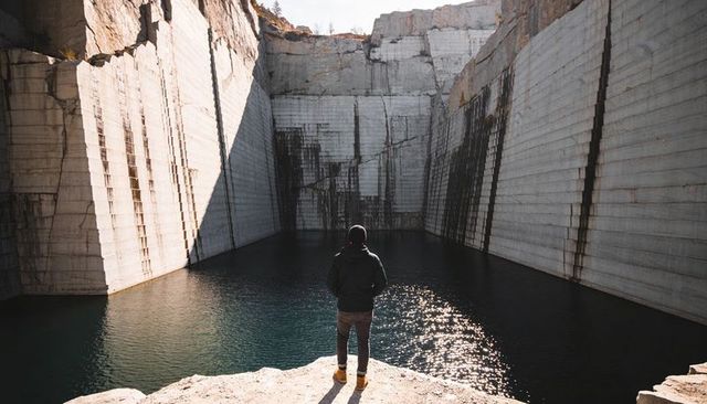 Hiker standing on ledge facing reflective quarry pool surrounded by towering stone walls