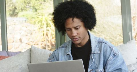 Remote worker focusing on laptop in sunroom, denim jacket and nose ring, natural daylight