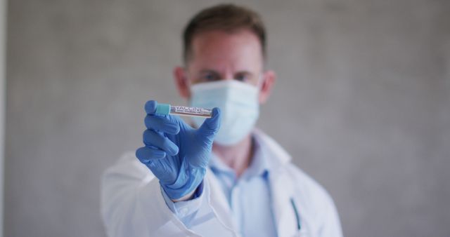 Doctor in lab coat, wearing protective mask and gloves, holding and displaying coronavirus test tube. Suitable for uses surrounding healthcare, COVID-19 awareness, pandemic information, medical research, laboratory safety, and public health campaigns.