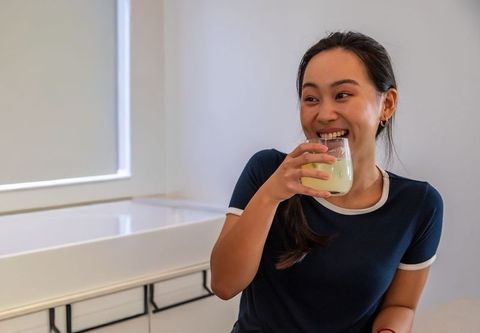 Asian woman smiling while holding glass of matcha latte at modern bright kitchen counter