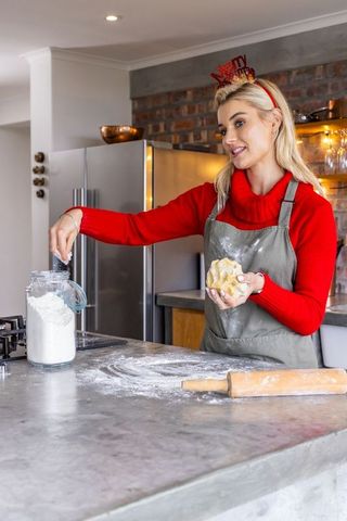 Woman wearing red sweater kneading dough while scooping flour in rustic kitchen