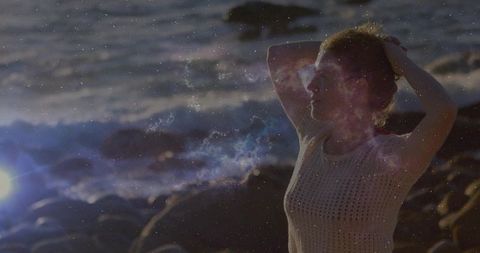 Standing woman holding hands behind head on rocky shore at sunset with cinematic backlight