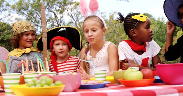 Children wearing playful costumes such as pirate hats and bunny ears are having fun at an outdoor celebration. The table is filled with healthy snacks and colorful bowls. Perfect for promoting children's parties, outdoor gatherings, creativity, healthy eating, and social interactions.