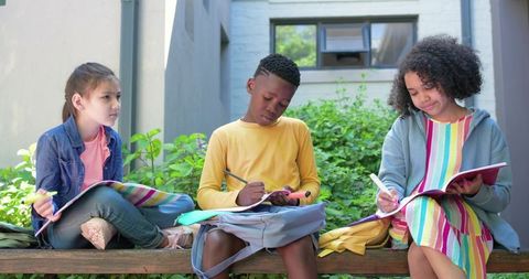 Children Studying Together on Outdoor Bench Writing in Notebooks Sharing Backpacks