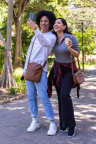 Sunlit park selfie by diverse couple smiling and walking with coffee and crossbody bags