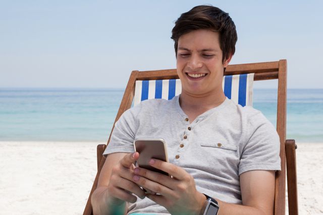 Smiling man sitting on sunlounger and using mobile phone on the beach