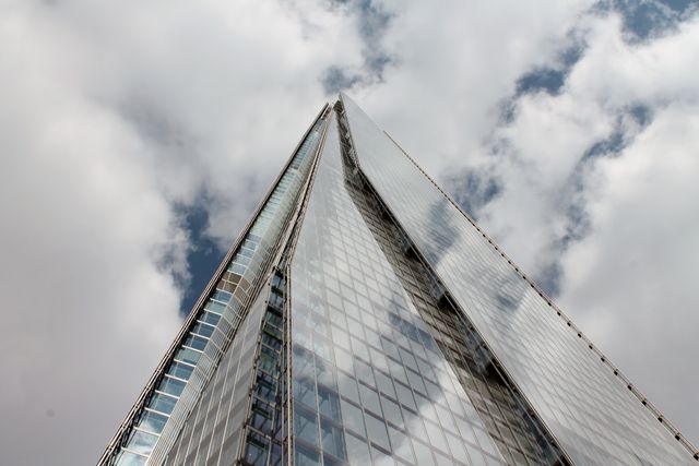 This image captures a low-angle view of a modern skyscraper with reflective glass surfaces against a cloudy sky backdrop. Useful for content related to urban development, business environments, modern architecture, city life, and construction projects.