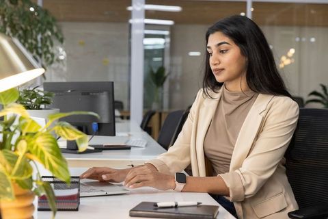 Indian businesswoman typing on laptop and checking smartwatch in modern open-plan office