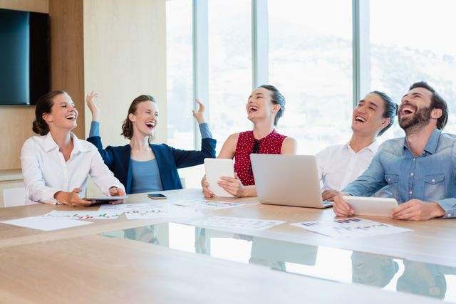 Business team members are gathered around a conference table, smiling and laughing during a meeting. They are using laptops and tablets, with charts and documents spread out on the table. This image is ideal for illustrating concepts of teamwork, collaboration, corporate culture, and a positive work environment. It can be used in business presentations, corporate websites, and marketing materials to convey a sense of productivity and employee satisfaction.