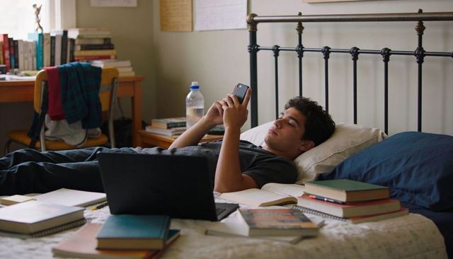 College student lying on bed using smartphone among books laptop and study materials