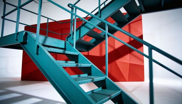 Turquoise metal staircase rising two flights with mid landing and red geometric panels