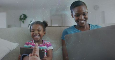 Smiling mother and daughter using laptop and tablet on couch, sharing digital moments