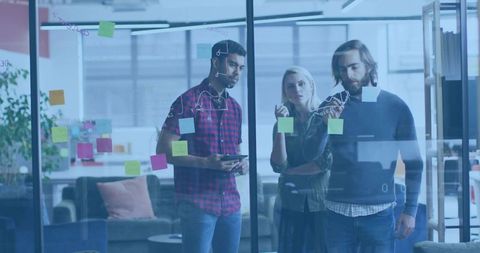 Collaborating team reviewing sticky notes and sketches on glass wall in modern lounge