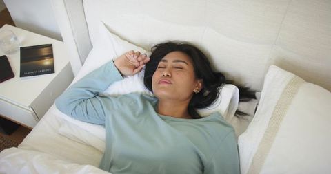 Mid-adult asian woman resting on bed in cozy bedroom wearing teal top with phone nearby