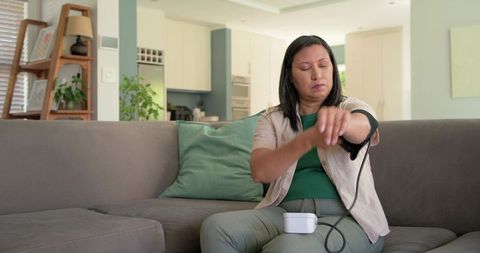 Middle-aged woman measuring blood pressure at home on couch with digital monitor