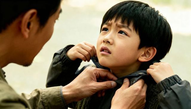 Father fastening son jacket zipper while boy looking up with worried expression outdoors
