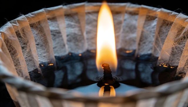 Macro burning wick in pleated paper lantern with dark oil fuel and warm amber flame glow