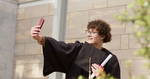 Young graduate taking selfie in cap and gown holding diploma on university campus