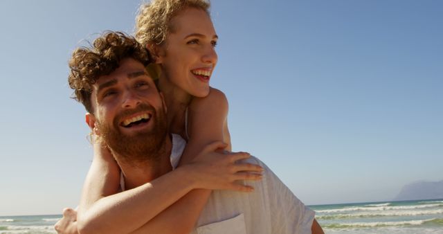 A young Caucasian couple enjoys a playful moment on a sunny beach, with copy space. Their joyful embrace and smiles suggest a carefree vacation or a romantic getaway.