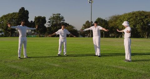 Four cricketers warming up on sunlit grass field wearing white uniforms and bucket hat