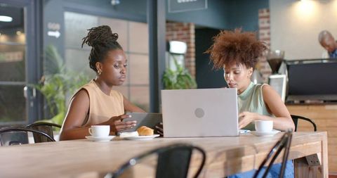 Black female colleagues collaborating over laptop and tablet in modern cafe coworking space