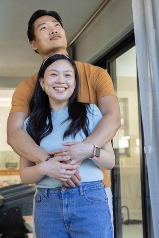 Asian couple embracing by sliding glass door, smiling, showing smartwatch and ring in sunlit home