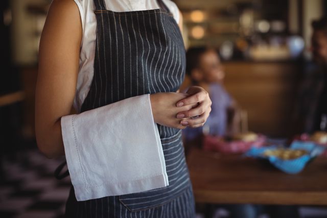 Waitress standing with a napkin in hand, wearing a striped apron in a restaurant. Ideal for use in articles or advertisements related to hospitality, restaurant service, dining experiences, and customer service training.