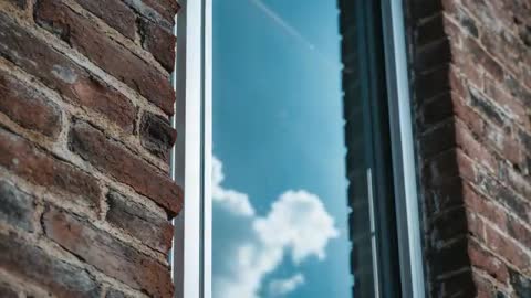 Camera Shifting Past Brick Facade Revealing Vertical Window Reflecting Clouds and Contrail