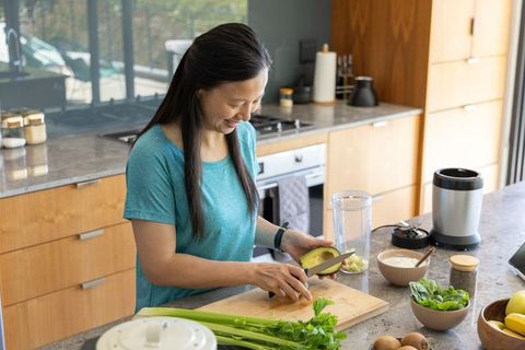 Asian woman slicing avocado at kitchen counter preparing green smoothie with blender