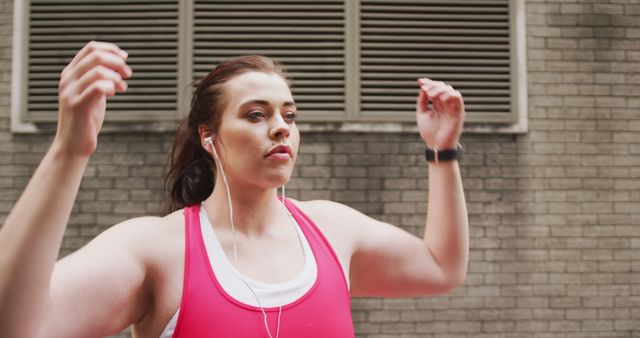 Young woman wearing earphones and fitness apparel exercising against a brick wall in an urban setting, portraying dedication and active lifestyle. Ideal for articles and advertisements focused on fitness, urban workouts, health promotions, and athletic motivation.